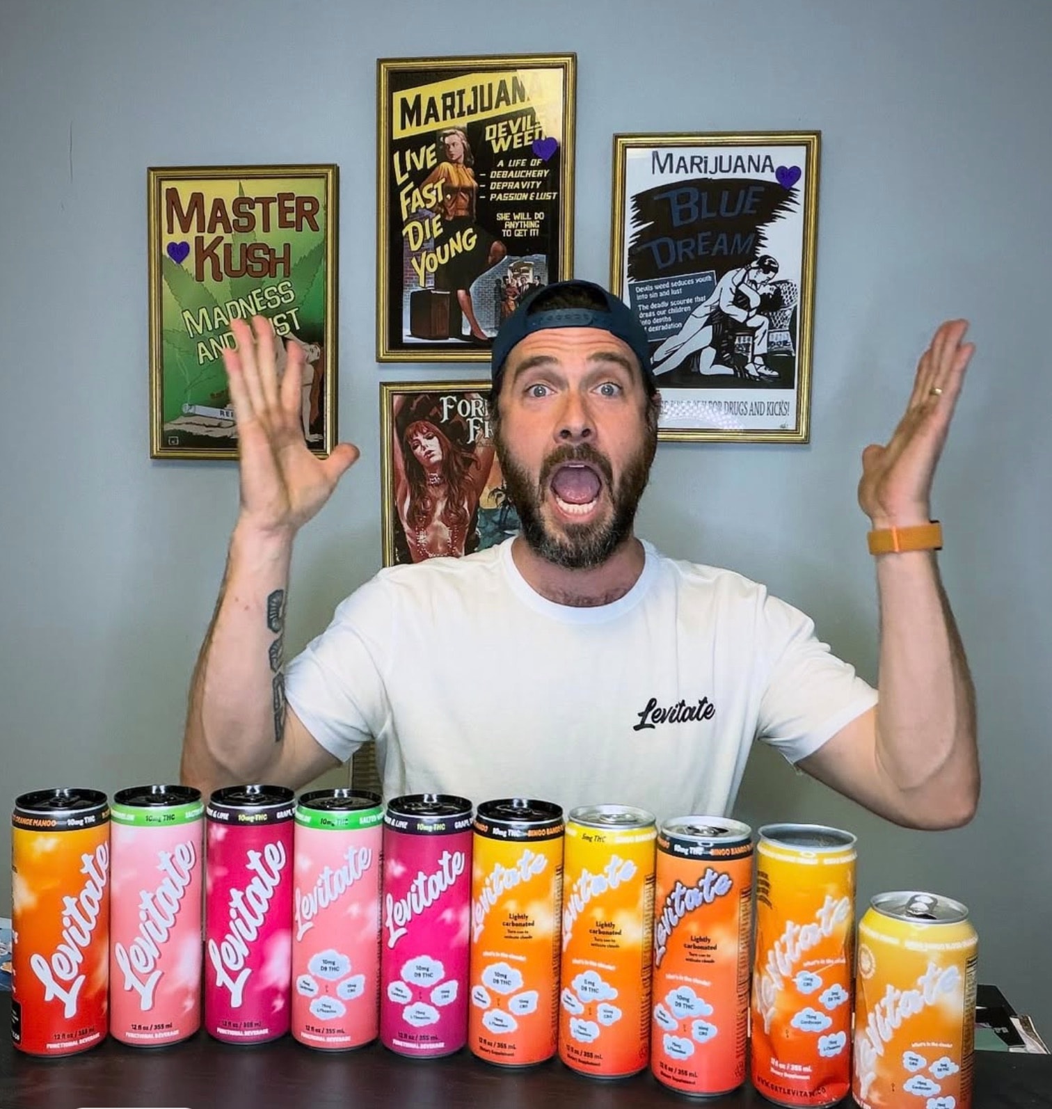 Albie Masland in a Levitate tee, arms up mid-cheer behind a lineup of every Levitate flavor, with vintage reefer-madness movie posters on the wall behind him
