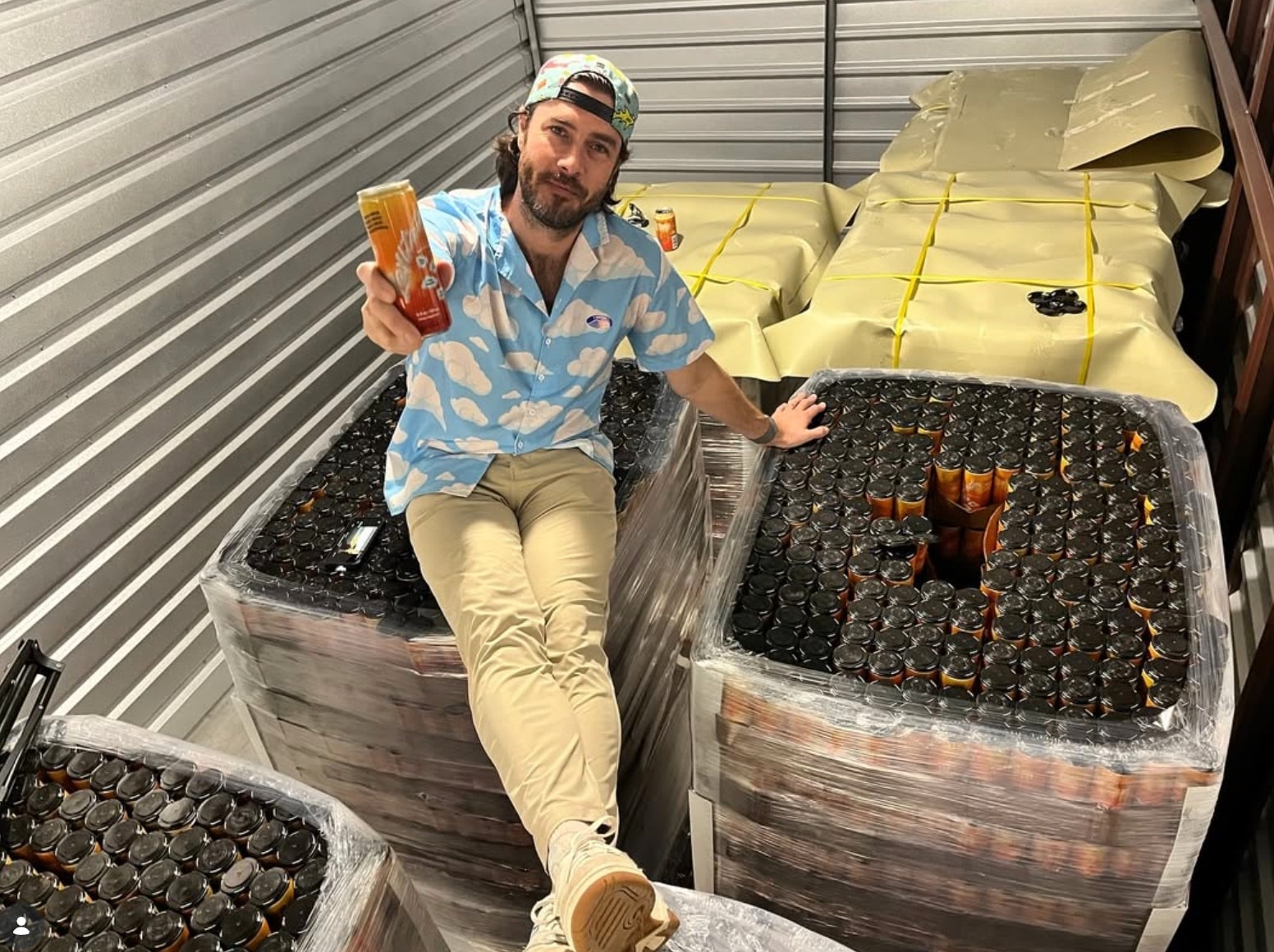 Albie Masland sitting on shrink-wrapped pallets of Levitate cans inside a warehouse, holding up a Blood Orange Mango