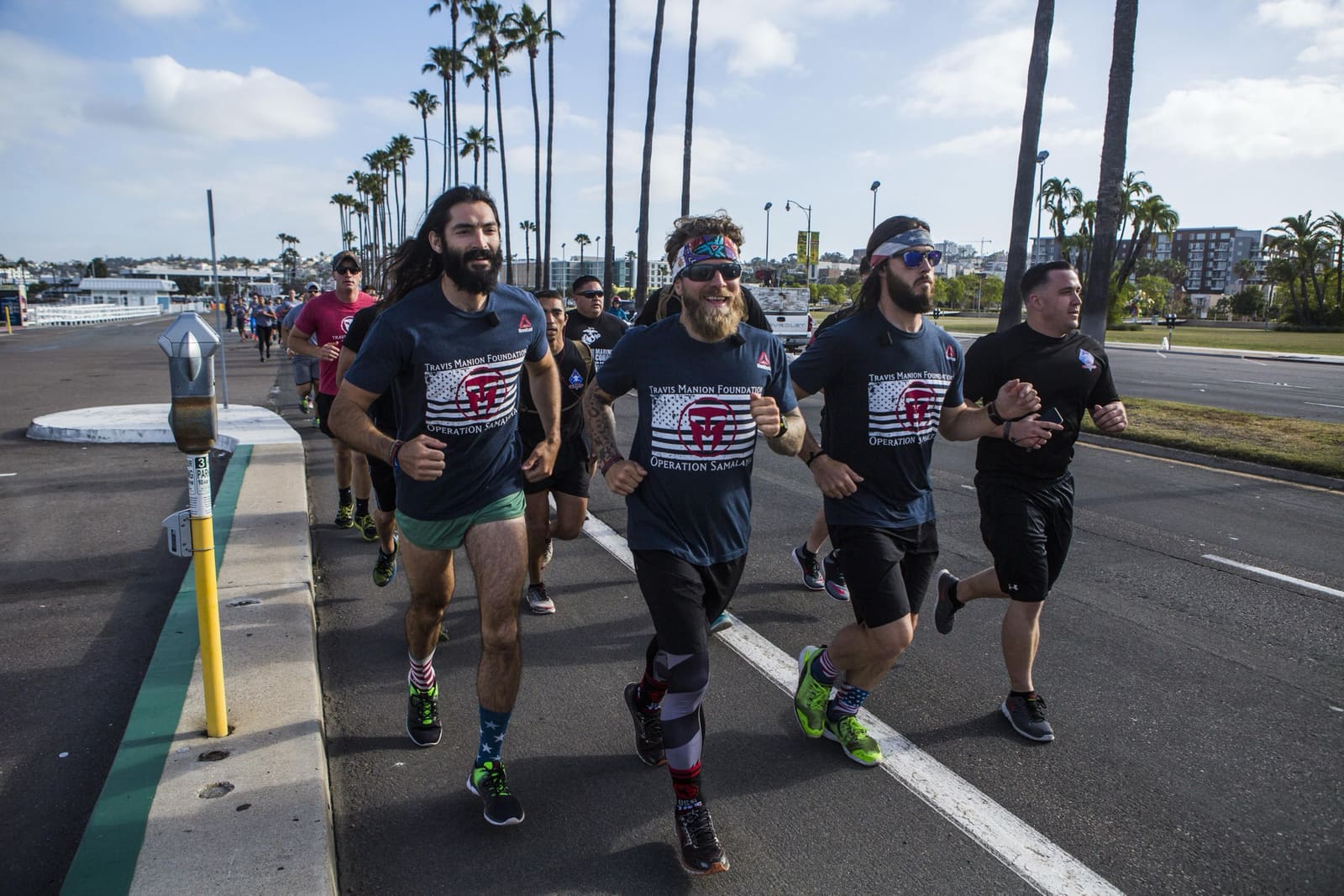Five runners in Travis Manion Foundation shirts mid-stride down a palm-lined San Diego boulevard, with the Pacific waterfront behind them